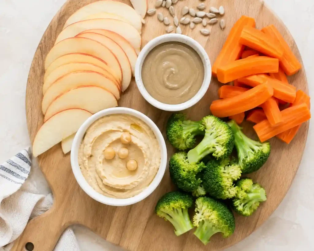 thin apple slices with a small dish of sunflower seed butter, steamed carrot and broccoli florets, and a small bowl of hummus